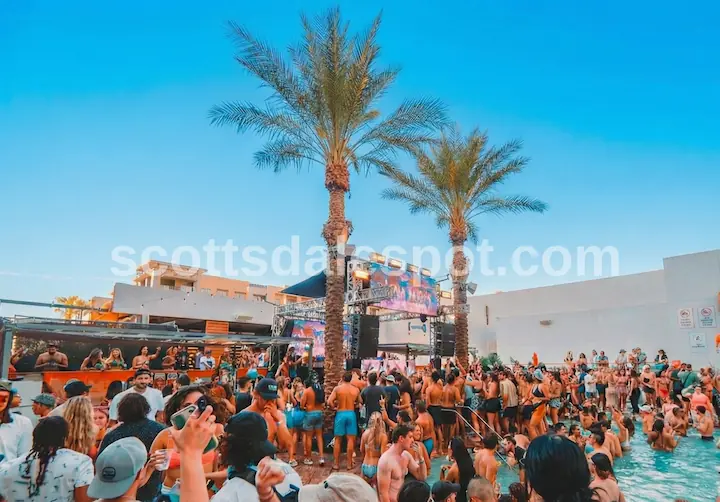 bachelorette party crowd celebrating at Maya Day + Nightclub pool party during sunset, featuring a DJ stage, palm trees, and golden hour